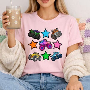 Może przedstawiać: Jasnor&oacute;żowa koszulka z kolorową grafiką monster truck&oacute;w i gwiazdami. Monster trucki są w r&oacute;żnych kolorach, w tym zielonym, pomarańczowym, niebieskim, ż&oacute;łtym i fioletowym. Gwiazdy są niebieskie, pomarańczowe, zielone i r&oacute;żowe.
