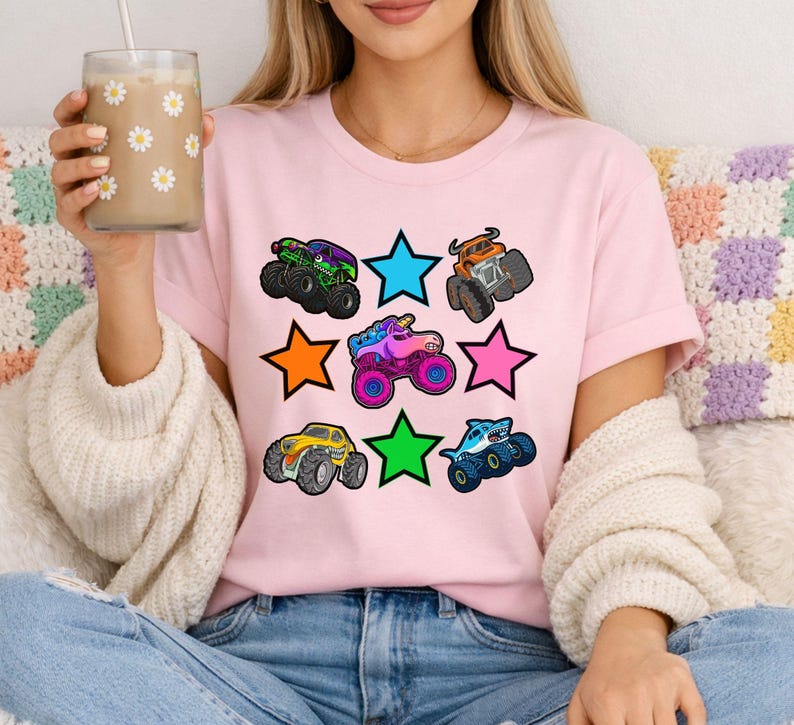 Może przedstawiać: Jasnor&oacute;żowa koszulka z kolorową grafiką monster truck&oacute;w i gwiazdami. Monster trucki są w r&oacute;żnych kolorach, w tym zielonym, pomarańczowym, niebieskim, ż&oacute;łtym i fioletowym. Gwiazdy są niebieskie, pomarańczowe, zielone i r&oacute;żowe.