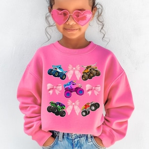 Op de afbeelding: Felroze sweatshirt met kleurrijke monstertruckafbeeldingen, waaronder een haai, een eenhoorn en een stier. Het ontwerp bevat roze strikken. De sweatshirt heeft een levendige roze kleur.