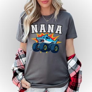 May include: Gray t-shirt with the word "NANA" in white block letters. Below the text is a cartoon shark monster truck in blue, white, and yellow. The shirt is worn with a red and black plaid shirt.