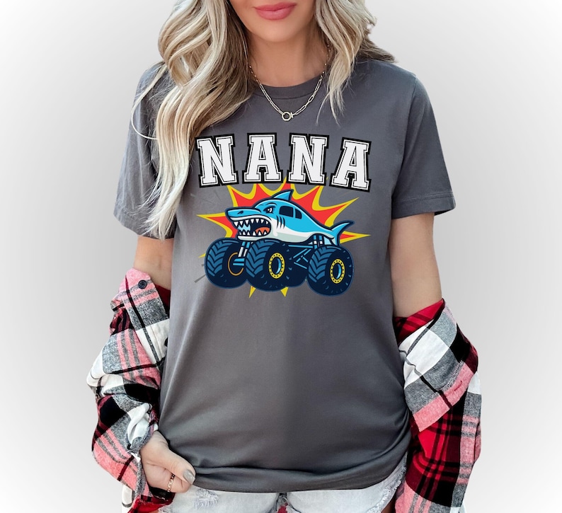 May include: Gray t-shirt with the word "NANA" in white block letters. Below the text is a cartoon shark monster truck in blue, white, and yellow. The shirt is worn with a red and black plaid shirt.