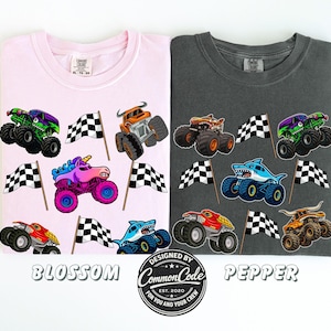 以下が含まれることがあります： ピンクと濃いグレーの2枚のTシャツが表示されています。ピンクのシャツには「Blossom」、グレーのシャツには「Pepper」と書かれています。どちらのシャツにも、カラフルなモンスタートラックのグラフィックとチェッカーフラッグが描かれています。