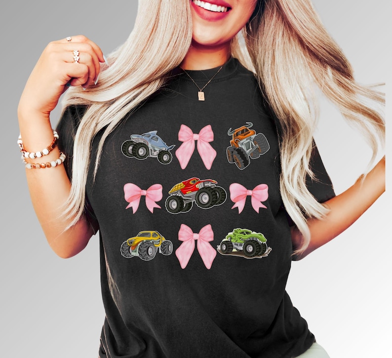 Lady Wearing a Black Comfort Colors t-shirt featuring monster trucks in various colors, including blue, orange, red, yellow, and green. Pink bows are interspersed between the truck in three rows. The shirt has a crew neck and short sleeves.