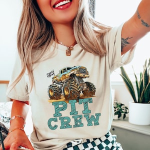 Pictured is a woman wearing a natural beige t-shirt featuring a colorful monster truck graphic with the words "PIT CREW" in teal. The monster truck has large tires and a checkered flag. The shirt is a casual style.