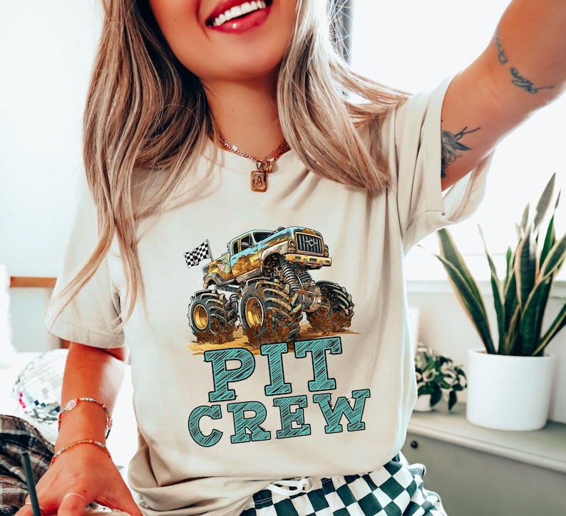 Pictured is a woman wearing a natural beige t-shirt featuring a colorful monster truck graphic with the words "PIT CREW" in teal. The monster truck has large tires and a checkered flag. The shirt is a casual style.