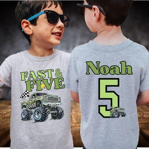Op de afbeelding: Een grijs t-shirt met de tekst "FAST & FIVE" en een monster truck afbeelding op de voorkant. De achterkant van het shirt toont de naam "Noah" en het nummer "5" in groen. Een kleine monster truck afbeelding staat onder het nummer.