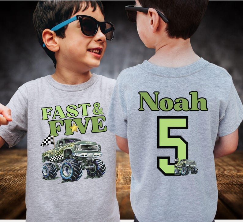Op de afbeelding: Een grijs t-shirt met de tekst "FAST & FIVE" en een monster truck afbeelding op de voorkant. De achterkant van het shirt toont de naam "Noah" en het nummer "5" in groen. Een kleine monster truck afbeelding staat onder het nummer.