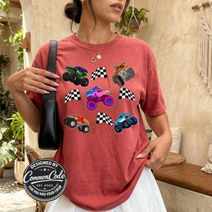 以下が含まれることがあります： レンガ色のTシャツ。クルーネックと半袖で、カラフルなモンスタートラックのグラフィックとチェッカーフラッグで飾られています。「DESIGNED BY COMMON CODE FOR YOU AND YOUR CREW EST. 2020」の文字が丸いパッチにプリントされています。