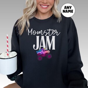 May include: Black sweatshirt with the words "Momster JAM" in white script and block letters. A colorful graphic of a unicorn monster truck is below the text. A white circle with the words "ANY NAME" is in the upper right corner.