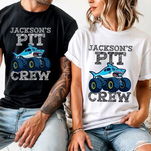 Op de afbeelding: Zwart en wit t-shirt en een wit t-shirt met de tekst "JACKSON'S PIT CREW". Elk shirt heeft een blauwe haaien monster truck afbeelding. De shirts zijn korte mouwen.
