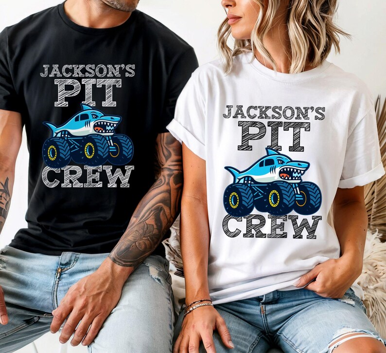 Op de afbeelding: Zwart en wit t-shirt en een wit t-shirt met de tekst "JACKSON'S PIT CREW". Elk shirt heeft een blauwe haaien monster truck afbeelding. De shirts zijn korte mouwen.