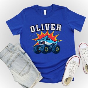 May include: A royal blue t-shirt featuring the name "OLIVER" in white block letters. Below the name is a cartoon shark on monster truck wheels, with a red and yellow burst background. Light blue denim shorts and white sneakers complete the look.