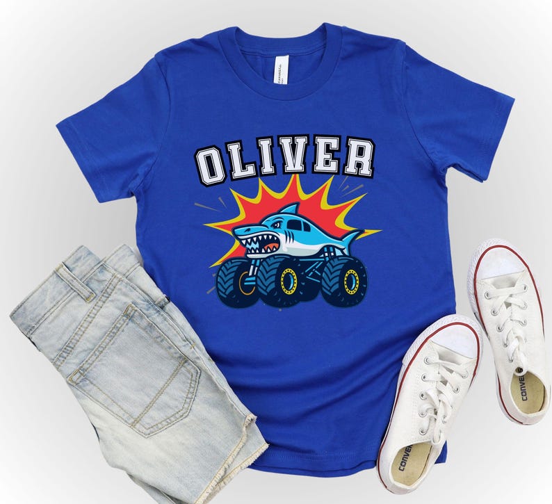 May include: A royal blue t-shirt featuring the name "OLIVER" in white block letters. Below the name is a cartoon shark on monster truck wheels, with a red and yellow burst background. Light blue denim shorts and white sneakers complete the look.