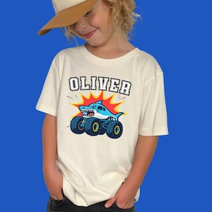 boy model is wearing a t-shirt in natural beige color that says "OLIVER" in varsity style letters in white with black outline in a arch - underneath is a shark monster truck cartoon style graphic. Perfect for Monster Truck Birthdays, Race Days, Birthday or Christmas Gift, Matching Family Shirts.