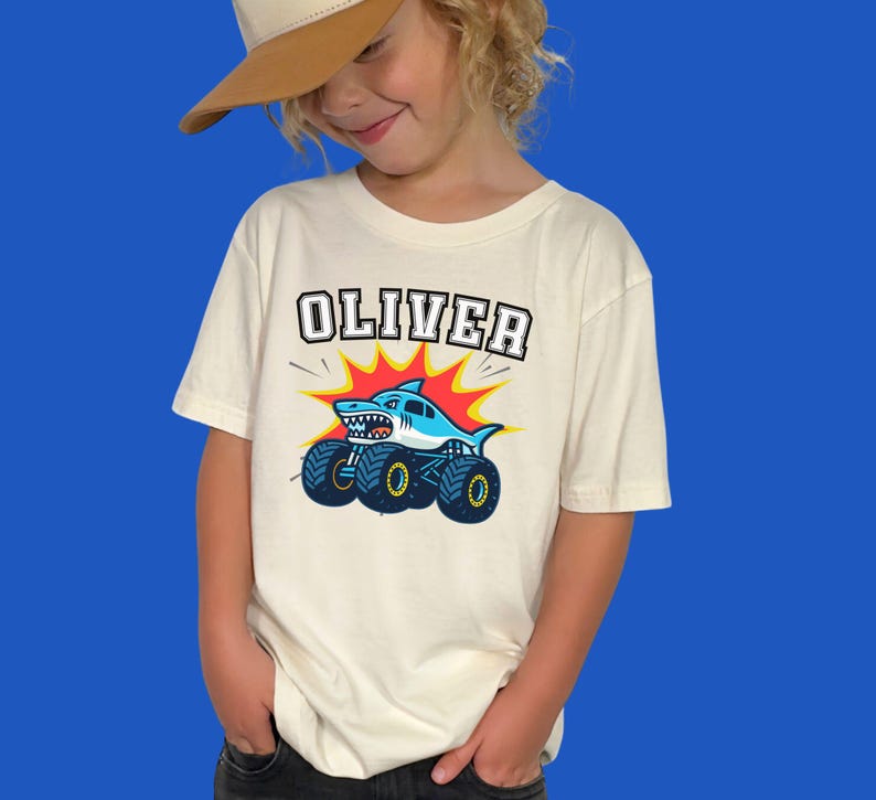 boy model is wearing a t-shirt in natural beige color that says "OLIVER" in varsity style letters in white with black outline in a arch - underneath is a shark monster truck cartoon style graphic. Perfect for Monster Truck Birthdays, Race Days, Birthday or Christmas Gift, Matching Family Shirts.