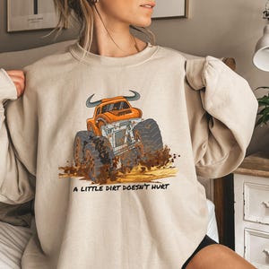Op de afbeelding: Beige sweatshirt met een cartoon monstertruck in oranje en wit, met zwarte banden en modderspatten. De tekst "A LITTLE DIRT DOESN'T HURT" staat eronder gedrukt. De sweatshirt heeft lange mouwen.