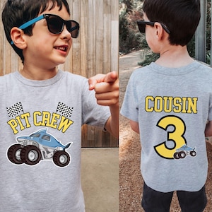 May include: A gray t-shirt with a shark monster truck graphic and the words "PIT CREW" in yellow. The back of the shirt says "COUSIN" and the number "3" in yellow. A small monster truck graphic is below the number.