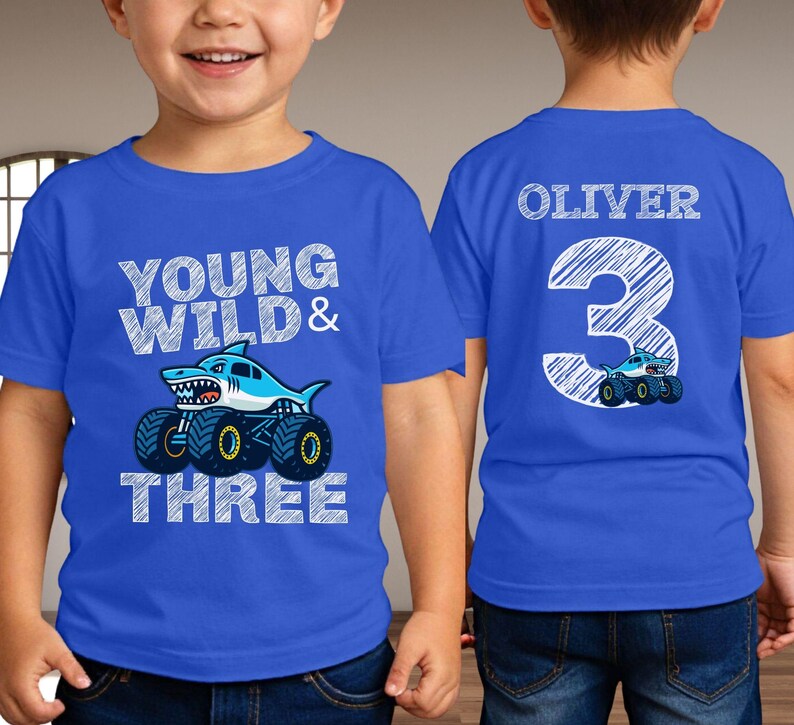 Op de afbeelding: Blauw T-shirt met een haaienmonster truck ontwerp. De voorkant zegt "Young Wild & Three". De achterkant toont de naam "Oliver" en het nummer "3" met een monstertruck.