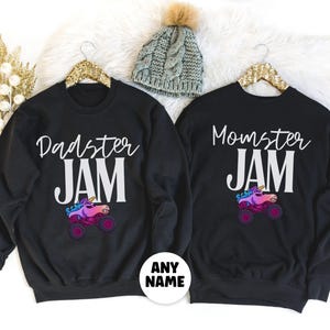 May include: Black sweatshirts with the phrases "Dadster JAM" and "Momster JAM" in white script. Each shirt features a colorful monster truck with a unicorn horn. A gray knit beanie with a pom-pom is also visible.