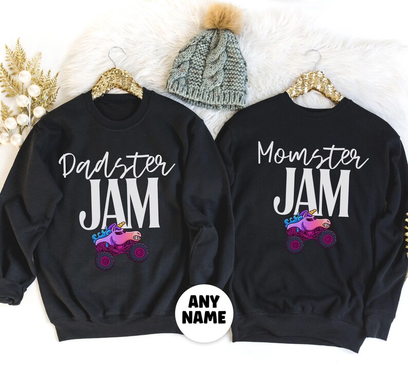 May include: Black sweatshirts with the phrases "Dadster JAM" and "Momster JAM" in white script. Each shirt features a colorful monster truck with a unicorn horn. A gray knit beanie with a pom-pom is also visible.