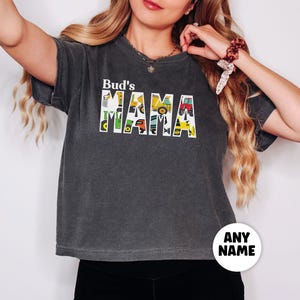 May include: Dark gray t-shirt with the text "Bud's MAMA" in white block letters. The letters are filled with colorful illustrations of construction vehicles. A white circle with the text "ANY NAME" is at the bottom.