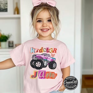 Puede incluir: Camiseta rosa claro con un gr&aacute;fico de cami&oacute;n monstruo colorido y las palabras "Kidster Jam". El dise&ntilde;o incluye un cami&oacute;n rosa con ruedas negras y una bandera a cuadros.