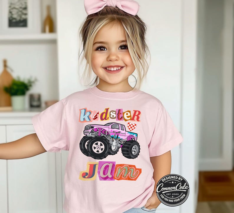 Puede incluir: Camiseta rosa claro con un gr&aacute;fico de cami&oacute;n monstruo colorido y las palabras "Kidster Jam". El dise&ntilde;o incluye un cami&oacute;n rosa con ruedas negras y una bandera a cuadros.
