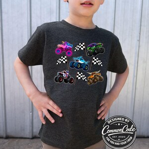 May include: A dark gray t-shirt featuring colorful monster truck graphics and checkered flags. The trucks have unique designs, including a shark, a unicorn, and a bull. The shirt has a round logo that says "Designed by Common Code Est. 2020 For You and Your Crew."