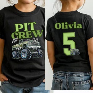 Op de afbeelding: Zwart T-shirt met een groene "PIT CREW" opdruk en een monster truck illustratie. De achterkant van het shirt toont de naam "Olivia", het nummer "5" en een monster truck afbeelding. Het shirt wordt door een kind gedragen.