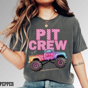 Pode incluir: T-shirt cinza escuro com as palavras "PIT CREW" em letras rosa xadrez. Um caminh&atilde;o monstro colorido com um la&ccedil;o rosa &eacute; apresentado abaixo do texto. A camisa &eacute; um estilo confort&aacute;vel e casual.