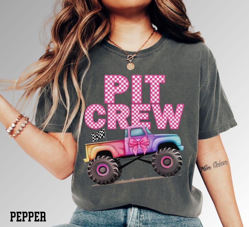 Pode incluir: T-shirt cinza escuro com as palavras "PIT CREW" em letras rosa xadrez. Um caminh&atilde;o monstro colorido com um la&ccedil;o rosa &eacute; apresentado abaixo do texto. A camisa &eacute; um estilo confort&aacute;vel e casual.