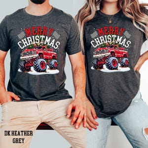 May include: Dark heather grey t-shirts with a red monster truck graphic and the text "MERRY CHRISTMAS". The truck has large tires and is set against a snowy background. The text "DK HEATHER GREY" is at the bottom.