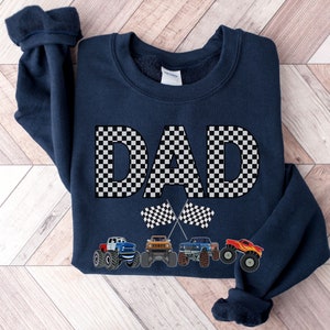 Op de afbeelding: Marineblauw sweatshirt met het woord 'DAD' in een zwart-wit dambordpatroon. Daaronder staan twee geblokte vlaggen en een rij kleurrijke monstertrucks.