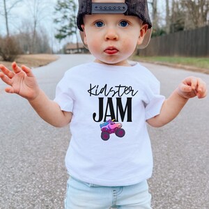 May include: A white t-shirt with the text "Kidster JAM" in black script and a colorful monster truck graphic. The shirt is worn by a young child wearing a black and tan baseball cap and light blue jeans.