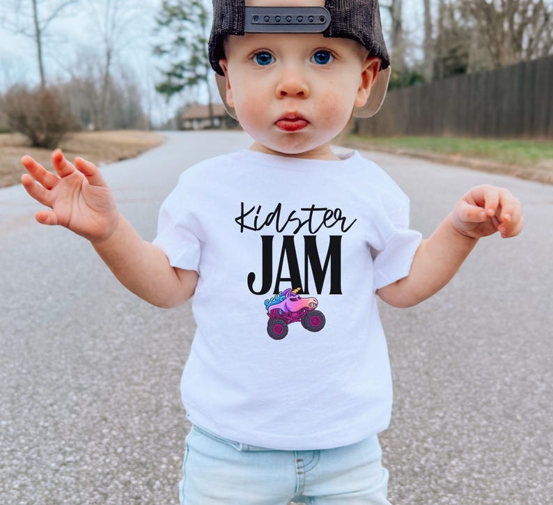 May include: A white t-shirt with the text "Kidster JAM" in black script and a colorful monster truck graphic. The shirt is worn by a young child wearing a black and tan baseball cap and light blue jeans.