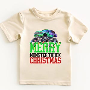 Puede incluir: Una camiseta beige de manga corta con un gr&aacute;fico de cami&oacute;n monstruo verde y el texto "Merry Monster Truck Christmas" en verde y rojo.