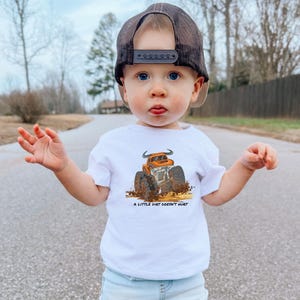 May include: A white t-shirt with an orange monster truck graphic and the words "A LITTLE DIRT DOESN'T HURT." The child is wearing a black mesh trucker cap. The image is taken outdoors.