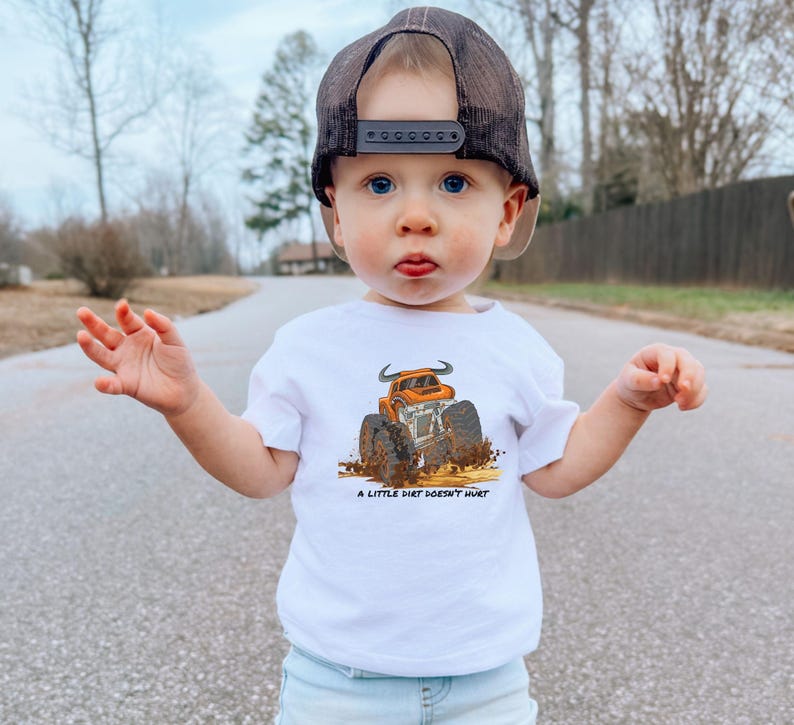 May include: A white t-shirt with an orange monster truck graphic and the words "A LITTLE DIRT DOESN'T HURT." The child is wearing a black mesh trucker cap. The image is taken outdoors.
