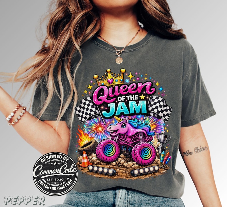 woman is wearing a unisex comfort colors tee in Pepper with a Pink Sparkle Unicorn Smash Truck that says "Queen of the Jam" in bubble letters with racing checkered flags, fireworks, stars, and a tire with fire. Very Fun Vibrant Monster truck family Matching shirt is perfect for Monster truck fans and Race Day Outfits. Mommy and Mini Matching outfits, Boy Mama Shirt. Matching Daughter and Mother outfit for Car shows and truck racing events, Pit Party, Birthday Party shirt with Monster truck theme