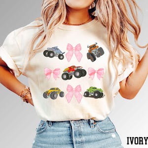 K&ouml;nnte beinhalten: Elfenbeinfarbenes T-Shirt mit einem Design aus sechs bunten Monstertrucks und rosa Schleifen. Die Monstertrucks sind in verschiedenen Farben, darunter Blau, Orange, Rot, Gelb und Gr&uuml;n. Das Wort "IVORY" ist unten aufgedruckt.