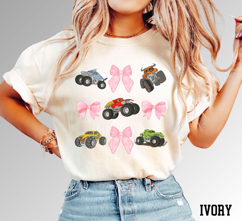 K&ouml;nnte beinhalten: Elfenbeinfarbenes T-Shirt mit einem Design aus sechs bunten Monstertrucks und rosa Schleifen. Die Monstertrucks sind in verschiedenen Farben, darunter Blau, Orange, Rot, Gelb und Gr&uuml;n. Das Wort "IVORY" ist unten aufgedruckt.