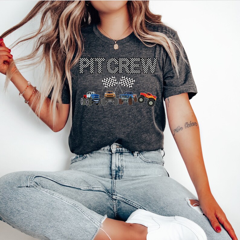 Puede incluir: Una camiseta gris con el texto "PIT CREW" en fuente de cuadros blancos y negros. La camiseta presenta una l&iacute;nea de camiones monstruo de dibujos animados con banderas a cuadros.