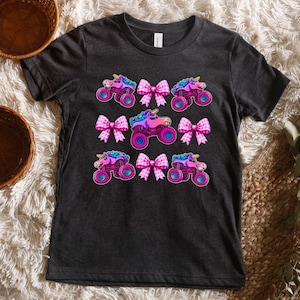 May include: Dark gray t-shirt featuring a colorful design of monster trucks with unicorn heads and pink bows. The trucks have blue, pink, and purple accents. The bows are pink and white.