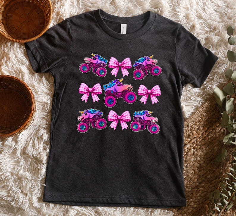 May include: Dark gray t-shirt featuring a colorful design of monster trucks with unicorn heads and pink bows. The trucks have blue, pink, and purple accents. The bows are pink and white.