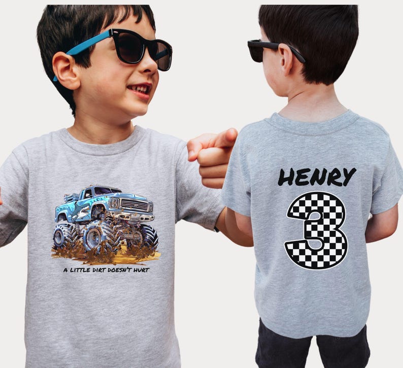 Peut inclure: Deux t-shirts gris. L'un pr&eacute;sente un dessin de camion monstre bleu avec le texte "A LITTLE DIRT DOESN'T HURT." L'autre t-shirt porte le nom "HENRY" et le chiffre "3" &agrave; damier au dos. Les deux t-shirts sont port&eacute;s par un enfant.