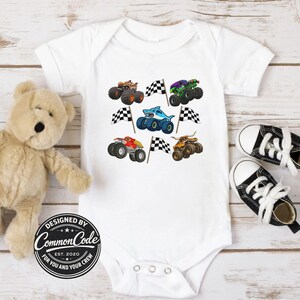 May include: White baby onesie featuring colorful monster truck designs and checkered flags. The onesie has a round neck and snap closures. The design includes a shark-themed monster truck. The text "DESIGNED BY COMMON CODE EST. 2020 FOR YOU AND YOUR CREW" is printed on the onesie.