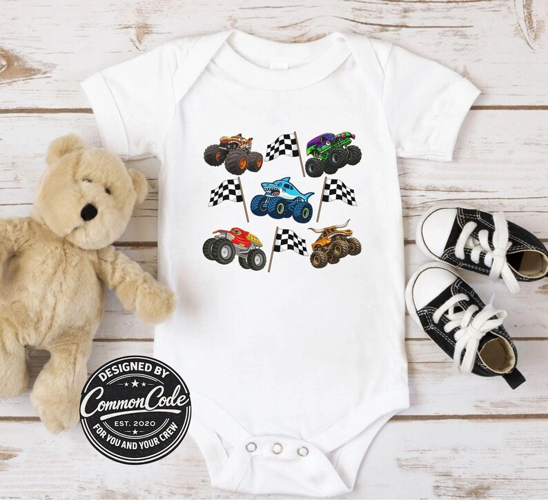 May include: White baby onesie featuring colorful monster truck designs and checkered flags. The onesie has a round neck and snap closures. The design includes a shark-themed monster truck. The text "DESIGNED BY COMMON CODE EST. 2020 FOR YOU AND YOUR CREW" is printed on the onesie.