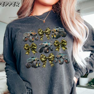 May include: Dark gray sweatshirt featuring a pattern of monster trucks and camouflage bows. The trucks are in various colors, with large tires. The word "PEPPER" is printed at the top left. A fun and playful design.