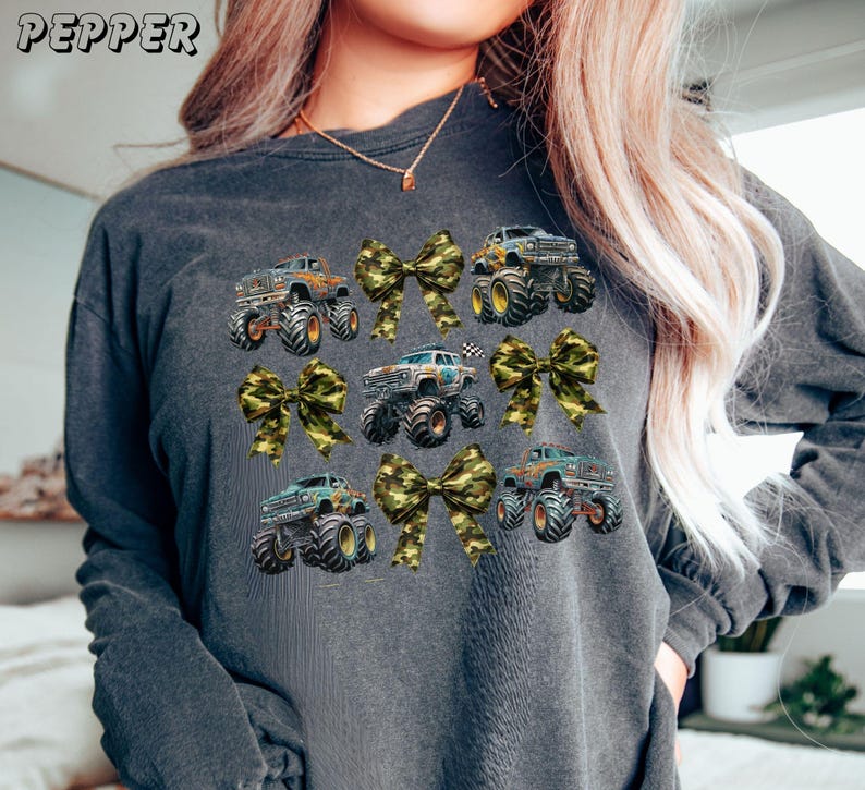 May include: Dark gray sweatshirt featuring a pattern of monster trucks and camouflage bows. The trucks are in various colors, with large tires. The word "PEPPER" is printed at the top left. A fun and playful design.