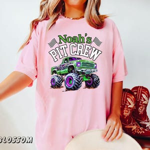 Pode incluir: Camiseta rosa claro com um desenho de um monster truck verde com rodas roxas e o texto "Noah's Pit Crew" em verde e branco. A camiseta tamb&eacute;m tem duas bandeiras quadriculadas.